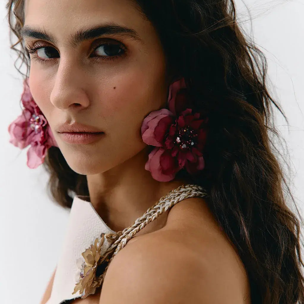 A close-up portrait of a woman wearing large, dark pink fabric flower earrings with jeweled centers and a matching braided floral shoulder detail on a white strap garment, against a plain light background.