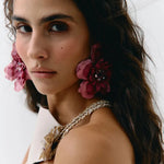 A close-up portrait of a woman wearing large, dark pink fabric flower earrings with jeweled centers and a matching braided floral shoulder detail on a white strap garment, against a plain light background.
