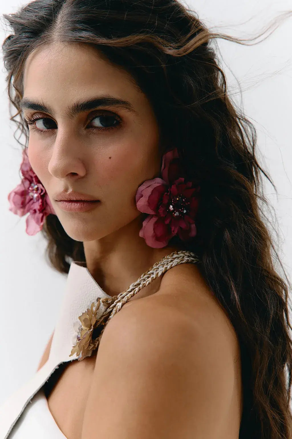 A close-up portrait of a woman wearing large, dark pink fabric flower earrings with jeweled centers and a matching braided floral shoulder detail on a white strap garment, against a plain light background.