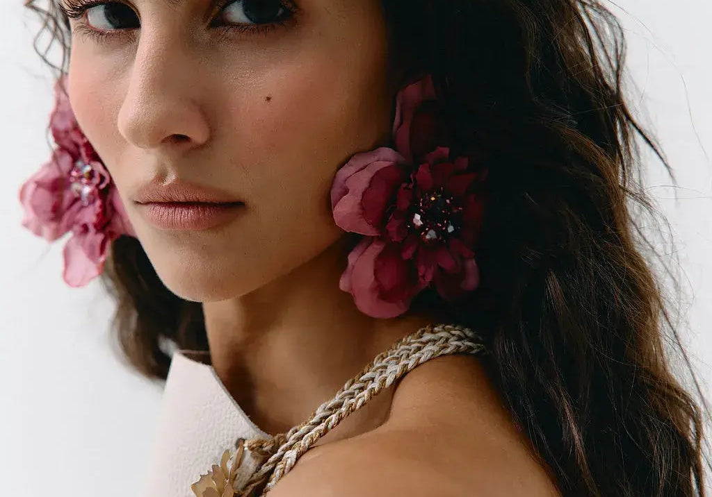A close-up portrait of a woman wearing large, dark pink fabric flower earrings with jeweled centers and a matching braided floral shoulder detail on a white strap garment, against a plain light background.
