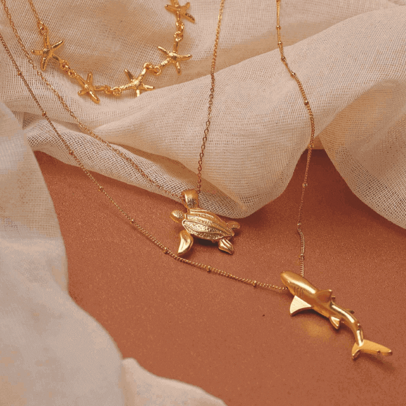 Gold necklaces with starfish, turtle and whale pendants on a beige fabric background.