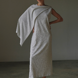 Woman wearing a textured white dress with a draped fabric over one shoulder against a neutral background.