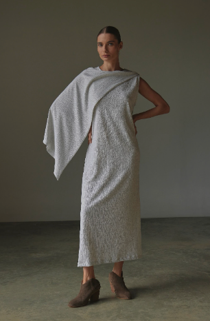 Woman wearing a textured white dress with a draped fabric over one shoulder against a neutral background.