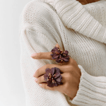 A close-up of a person wearing a chunky cream knit sweater and two large dark purple flower-shaped rings on their fingers.