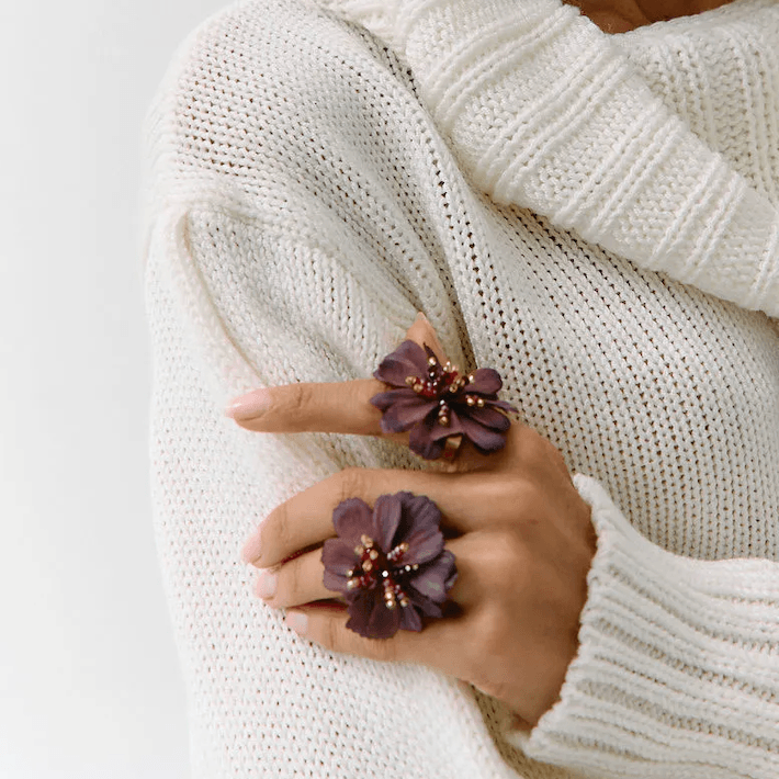 A close-up of a person wearing a chunky cream knit sweater and two large dark purple flower-shaped rings on their fingers.