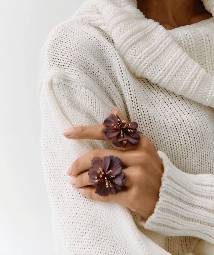 A close-up of a person wearing a chunky cream knit sweater and two large dark purple flower-shaped rings on their fingers.