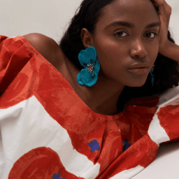 A woman wearing large, vibrant blue flower-shaped earrings and a red and white patterned top, posing against a neutral light beige background.