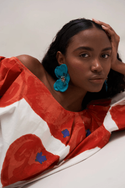A woman wearing large, vibrant blue flower-shaped earrings and a red and white patterned top, posing against a neutral light beige background.