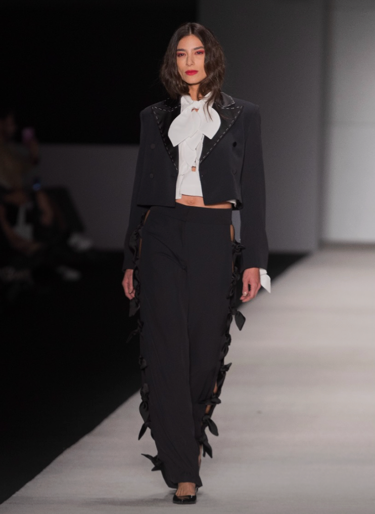 A model walks down a fashion runway wearing a black cropped jacket and matching wide-leg trousers with dramatic black ribbon or fabric ruffle accents along the edges, paired with a white high-neck bow blouse.