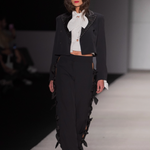 A model walks down a fashion runway wearing a black cropped jacket and matching wide-leg trousers with dramatic black ribbon or fabric ruffle accents along the edges, paired with a white high-neck bow blouse.