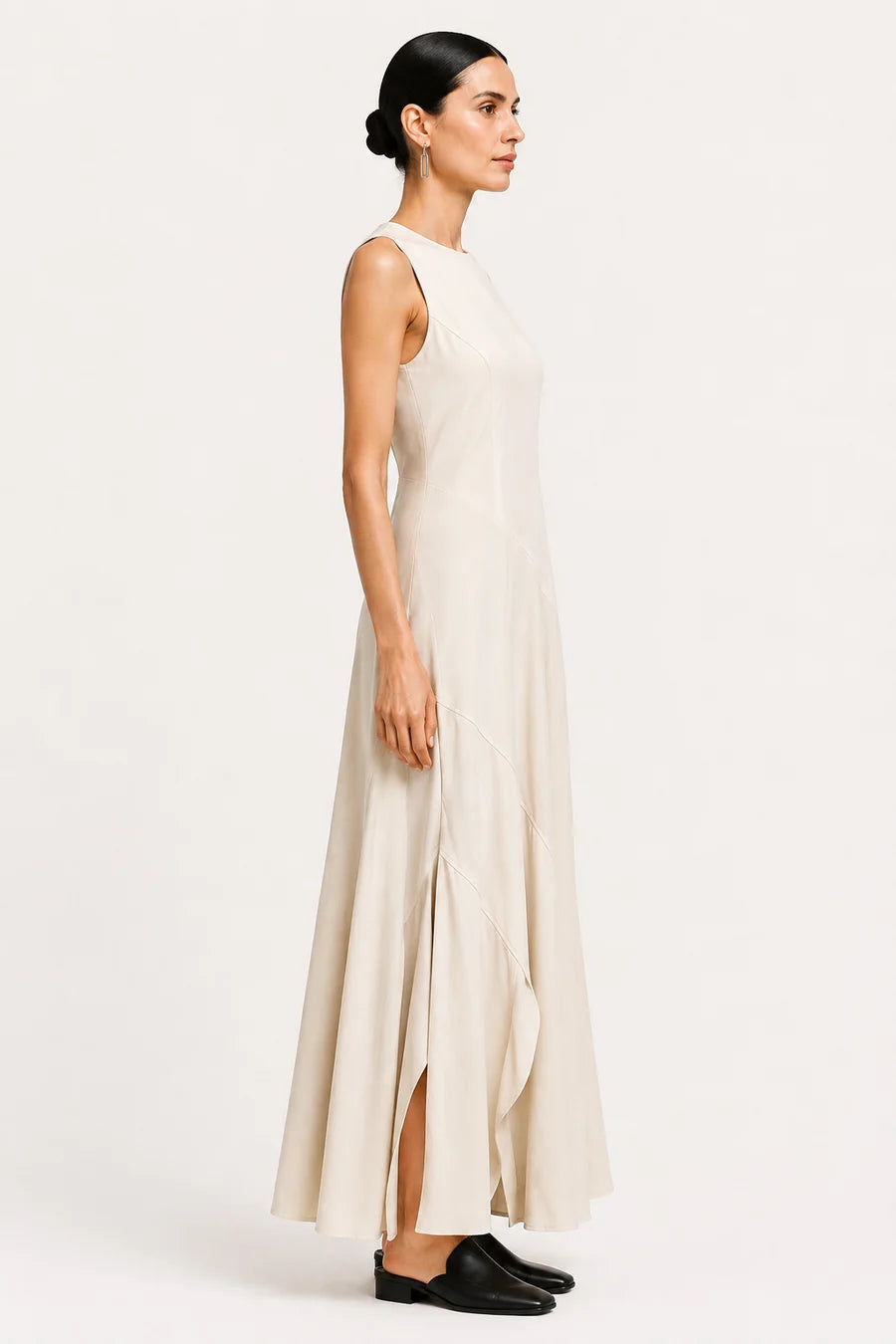 A woman modeling a sleeveless, floor-length cream dress with a high neckline and a side slit, paired with black flat shoes, against a plain white studio background.