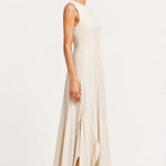 A woman modeling a sleeveless, floor-length cream dress with a high neckline and a side slit, paired with black flat shoes, against a plain white studio background.