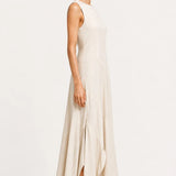 A woman modeling a sleeveless, floor-length cream dress with a high neckline and a side slit, paired with black flat shoes, against a plain white studio background.