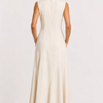 A woman viewed from the back wearing a sleeveless, floor-length cream dress with a straight neckline and a flared skirt, standing against a plain light background.