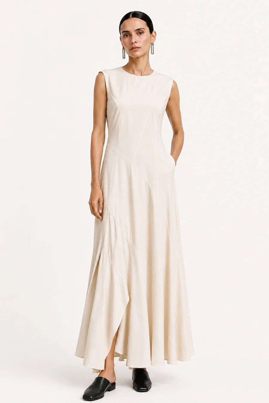 A woman modeling a sleeveless, floor-length cream dress with a high neckline and draped skirt, paired with black loafers, against a plain white studio background.