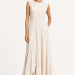 A woman modeling a sleeveless, floor-length cream dress with a high neckline and draped skirt, paired with black loafers, against a plain white studio background.