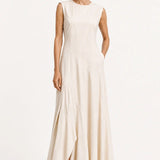 A woman modeling a sleeveless, floor-length cream dress with a high neckline and draped skirt, paired with black loafers, against a plain white studio background.