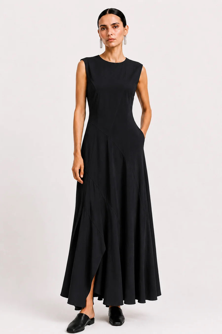A woman modeling a sleeveless black maxi dress with a round neckline, fitted bodice, and flowing skirt, standing against a plain light gray studio background.