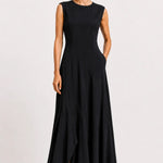 A woman modeling a sleeveless black maxi dress with a round neckline, fitted bodice, and flowing skirt, standing against a plain light gray studio background.