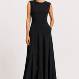 A woman modeling a sleeveless black maxi dress with a round neckline, fitted bodice, and flowing skirt, standing against a plain light gray studio background.