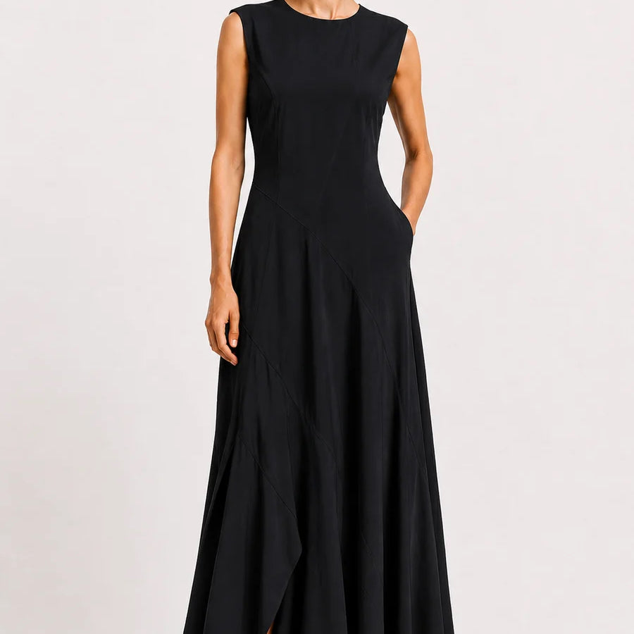 A woman modeling a sleeveless black maxi dress with a round neckline, fitted bodice, and flowing skirt, standing against a plain light gray studio background.
