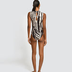 A woman stands with her back to the camera wearing a sleeveless one-piece swimsuit with a bold black and white zebra-like abstract pattern. She is photographed against a plain light gray studio background.