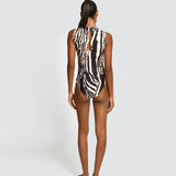 A woman stands with her back to the camera wearing a sleeveless one-piece swimsuit with a bold black and white zebra-like abstract pattern. She is photographed against a plain light gray studio background.