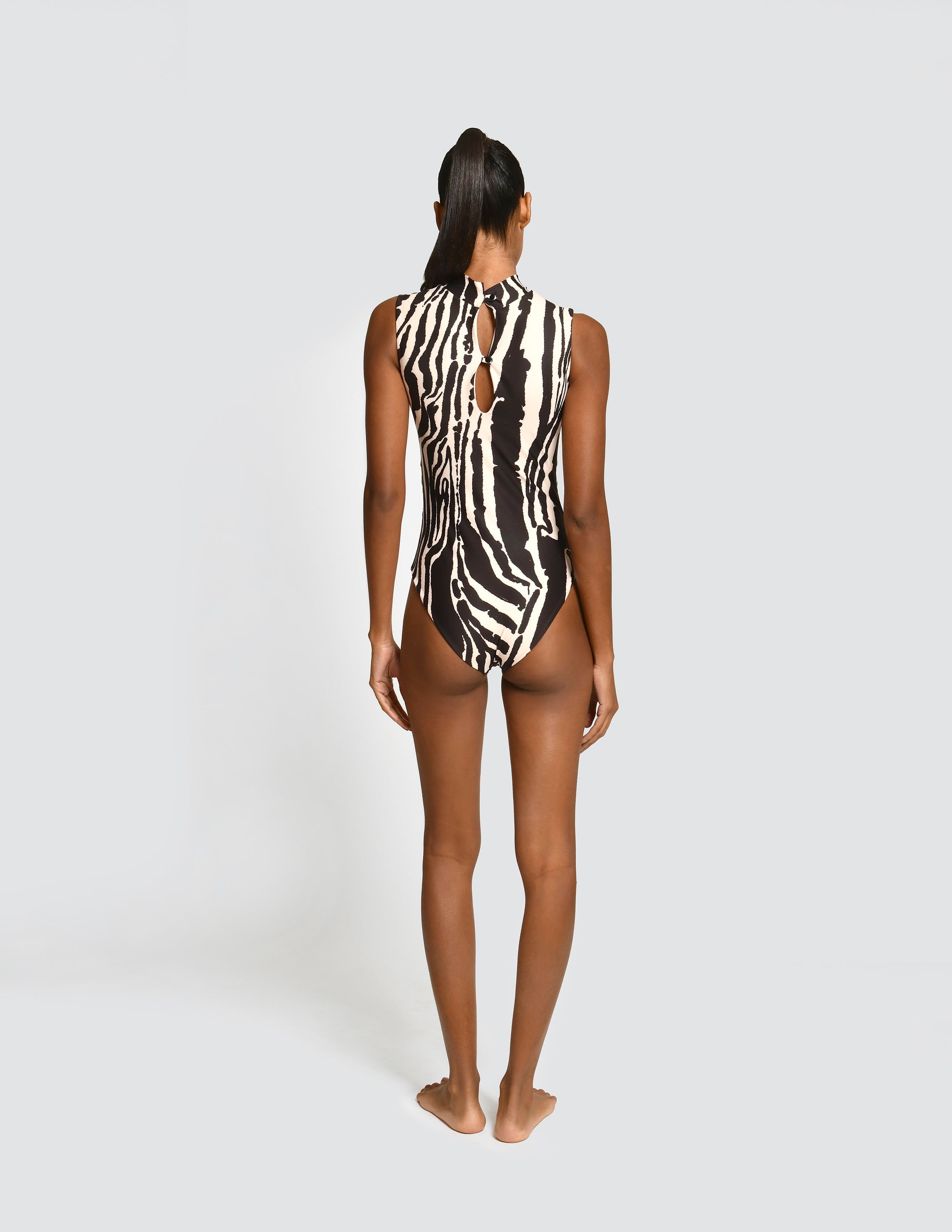 A woman stands with her back to the camera wearing a sleeveless one-piece swimsuit with a bold black and white zebra-like abstract pattern. She is photographed against a plain light gray studio background.