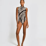 A model stands against a light gray studio background wearing a sleeveless one-piece swimsuit with a bold black and white zebra-like abstract pattern.