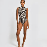A model stands against a light gray studio background wearing a sleeveless one-piece swimsuit with a bold black and white zebra-like abstract pattern.