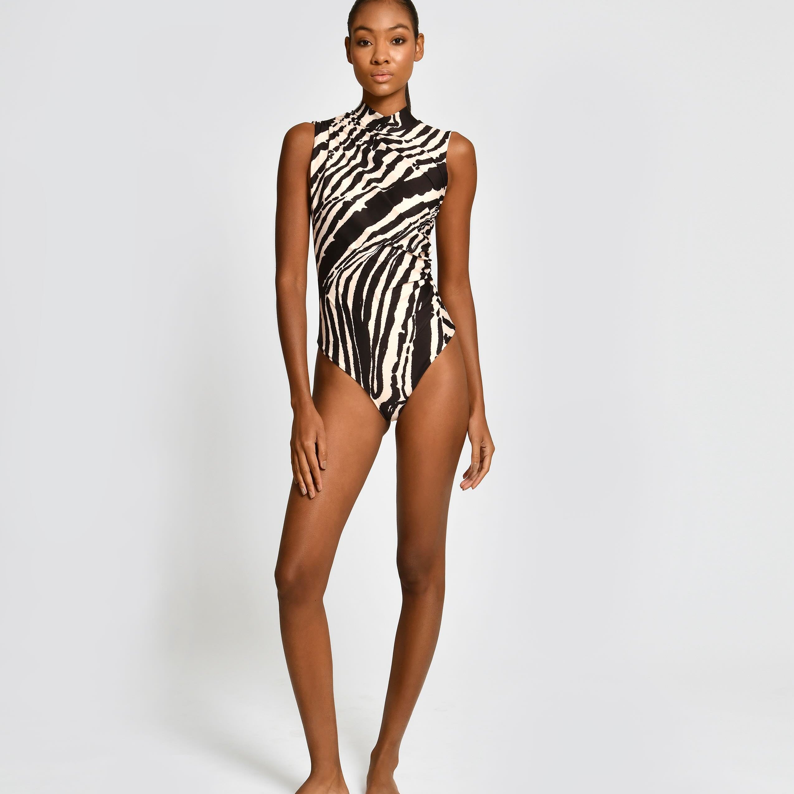 A model stands against a light gray studio background wearing a sleeveless one-piece swimsuit with a bold black and white zebra-like abstract pattern.