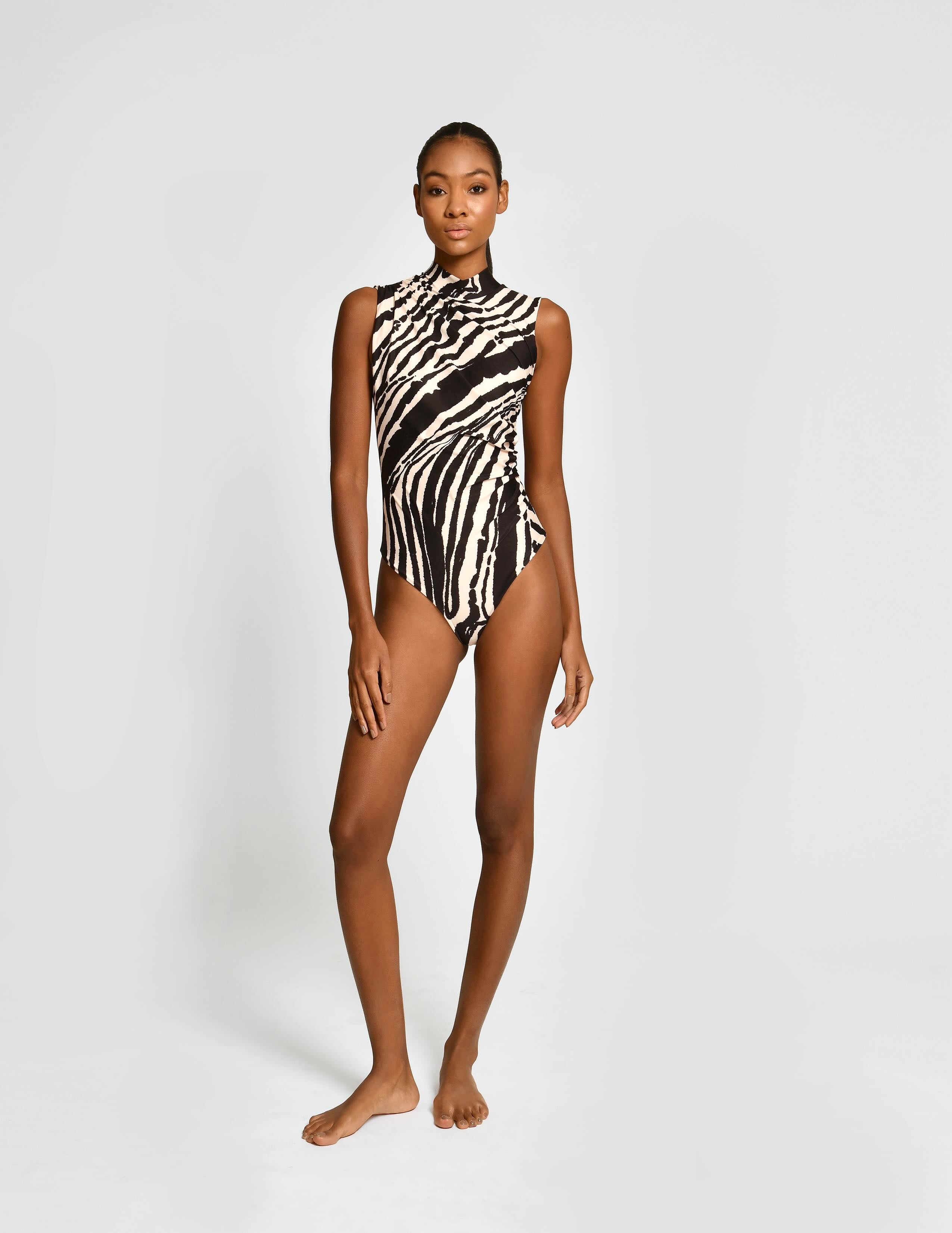 A model stands against a light gray studio background wearing a sleeveless one-piece swimsuit with a bold black and white zebra-like abstract pattern.