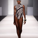 A model walks down a runway wearing a sleeveless black and white zebra-print one-piece swimsuit with strappy details on the upper arms, white sunglasses, and gold jewelry.