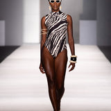 A model walks down a runway wearing a sleeveless black and white zebra-print one-piece swimsuit with strappy details on the upper arms, white sunglasses, and gold jewelry.