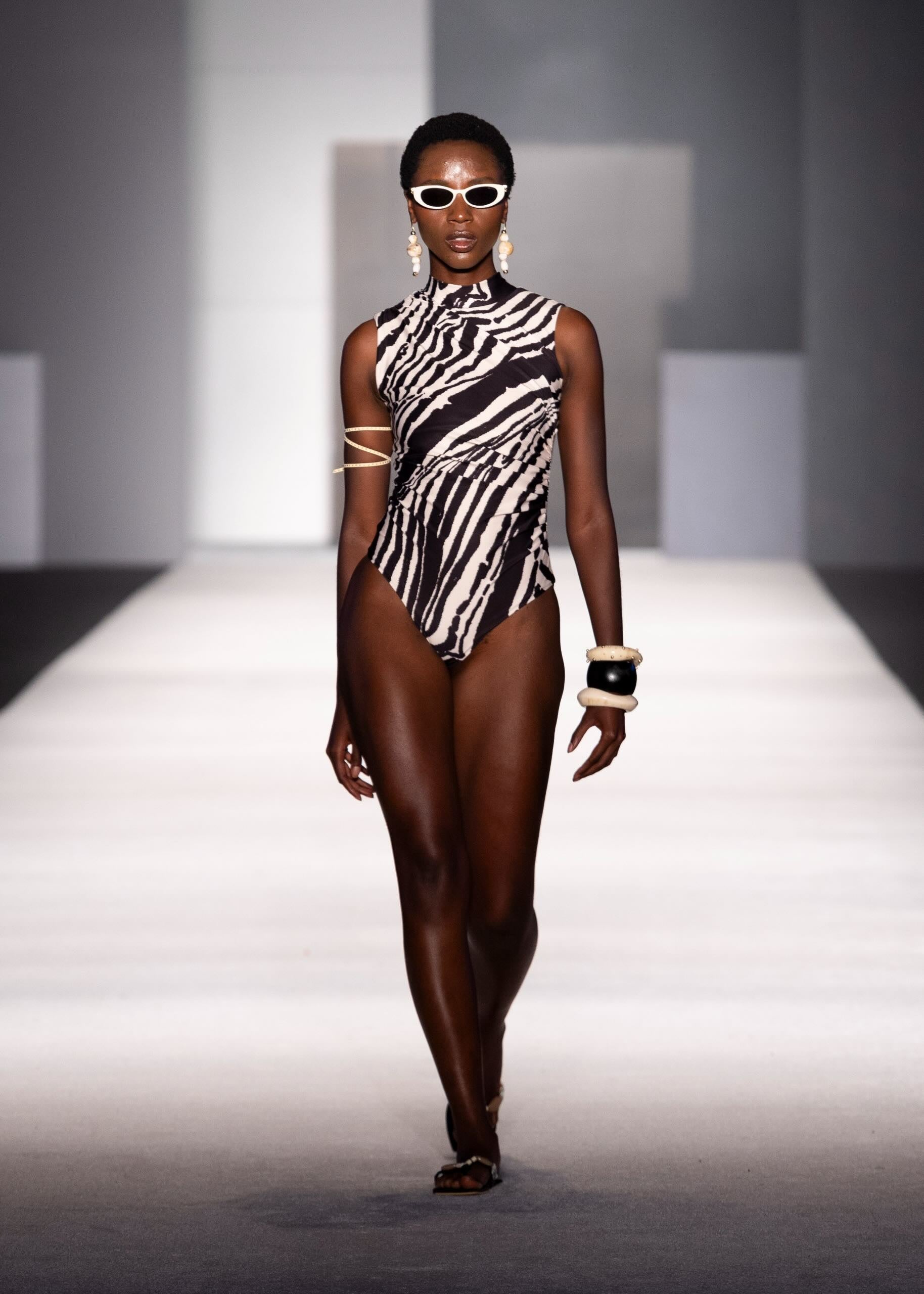 A model walks down a runway wearing a sleeveless black and white zebra-print one-piece swimsuit with strappy details on the upper arms, white sunglasses, and gold jewelry.