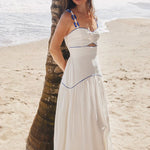 A woman wearing a white maxi dress with blue trim stands barefoot on a sandy beach next to a palm tree, with the ocean in the background.
