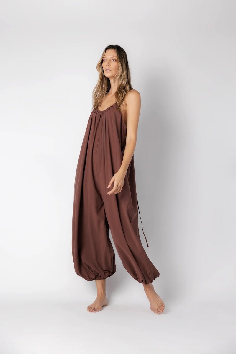 Model wearing a brown jumpsuit. She's standing behind a white background. The purpose of this image is to highlight the jumpsuit, which is available at EVAMAIA boutique in Los Angeles.