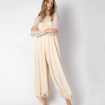 Model wearing a beige jumpsuit. She's standing behind a white background. The purpose of this image is to highlight the jumpsuit, which is available at EVAMAIA boutique in Los Angeles.