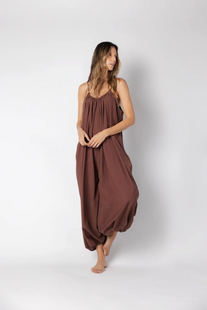 Model wearing a brown jumpsuit. She's standing behind a white background. The purpose of this image is to highlight the jumpsuit, which is available at EVAMAIA boutique in Los Angeles.