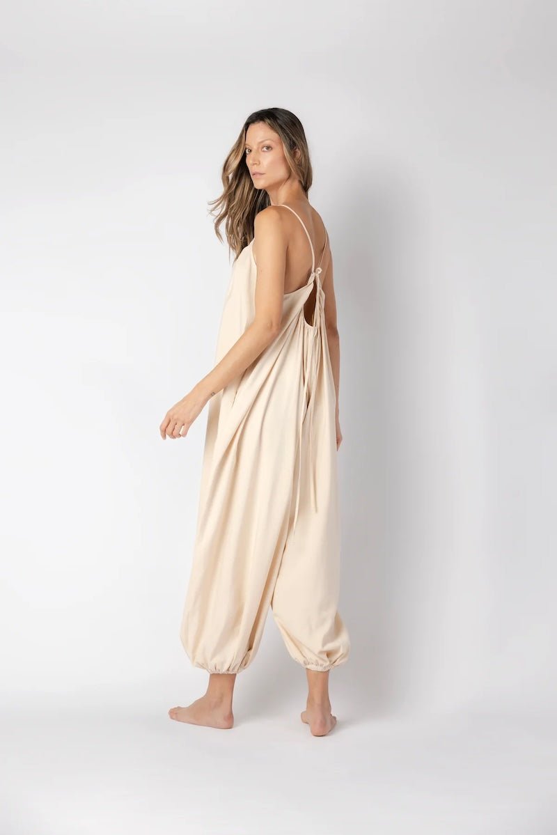 Model wearing a beige jumpsuit. She's standing behind a white background. The purpose of this image is to highlight the jumpsuit, which is available at EVAMAIA boutique in Los Angeles.