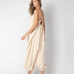 Model wearing a beige jumpsuit. She's standing behind a white background. The purpose of this image is to highlight the jumpsuit, which is available at EVAMAIA boutique in Los Angeles.