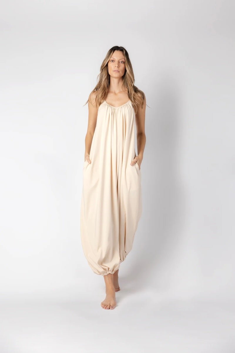 Model wearing a beige jumpsuit. She's standing behind a white background. The purpose of this image is to highlight the jumpsuit, which is available at EVAMAIA boutique in Los Angeles.