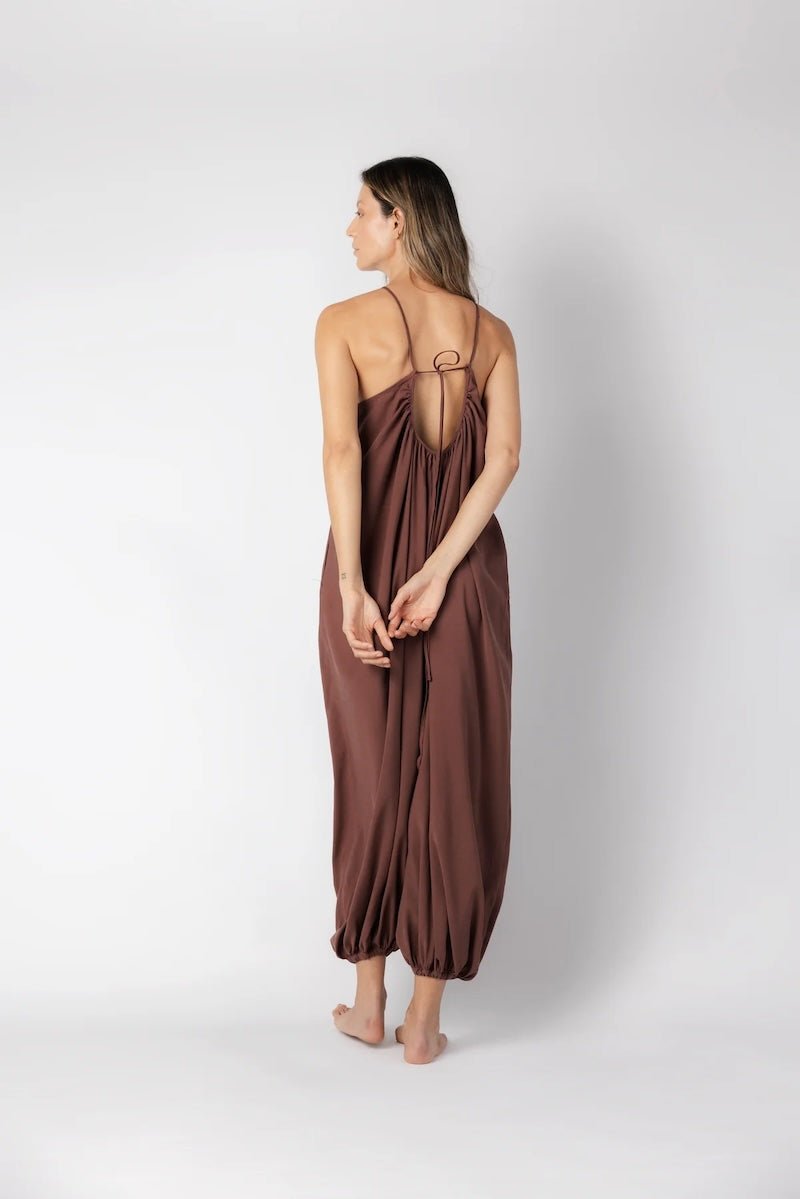 Model wearing a brown jumpsuit. She's standing behind a white background. The purpose of this image is to highlight the jumpsuit, which is available at EVAMAIA boutique in Los Angeles.