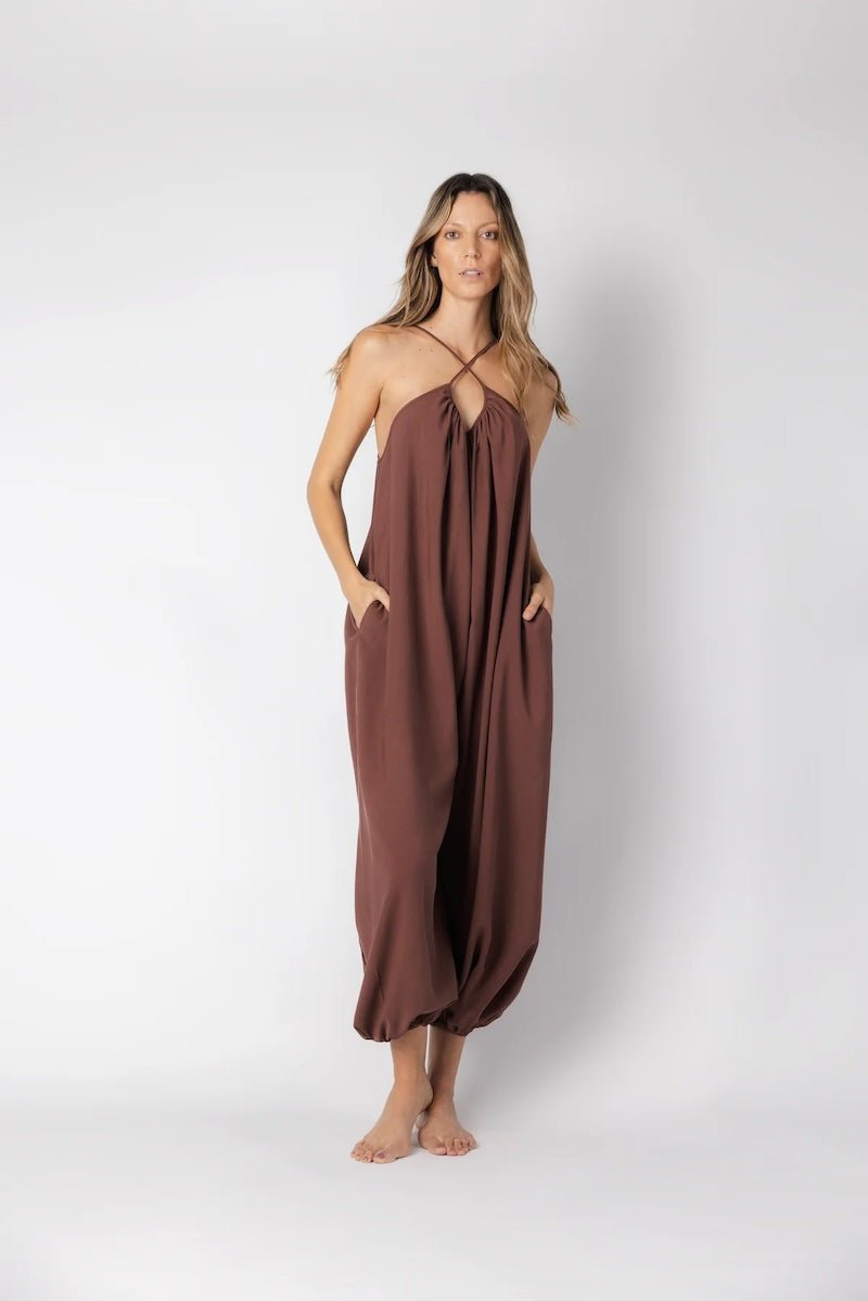 Model wearing a brown jumpsuit. She's standing behind a white background. The purpose of this image is to highlight the jumpsuit, which is available at EVAMAIA boutique in Los Angeles.