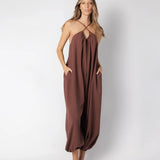 Model wearing a brown jumpsuit. She's standing behind a white background. The purpose of this image is to highlight the jumpsuit, which is available at EVAMAIA boutique in Los Angeles.