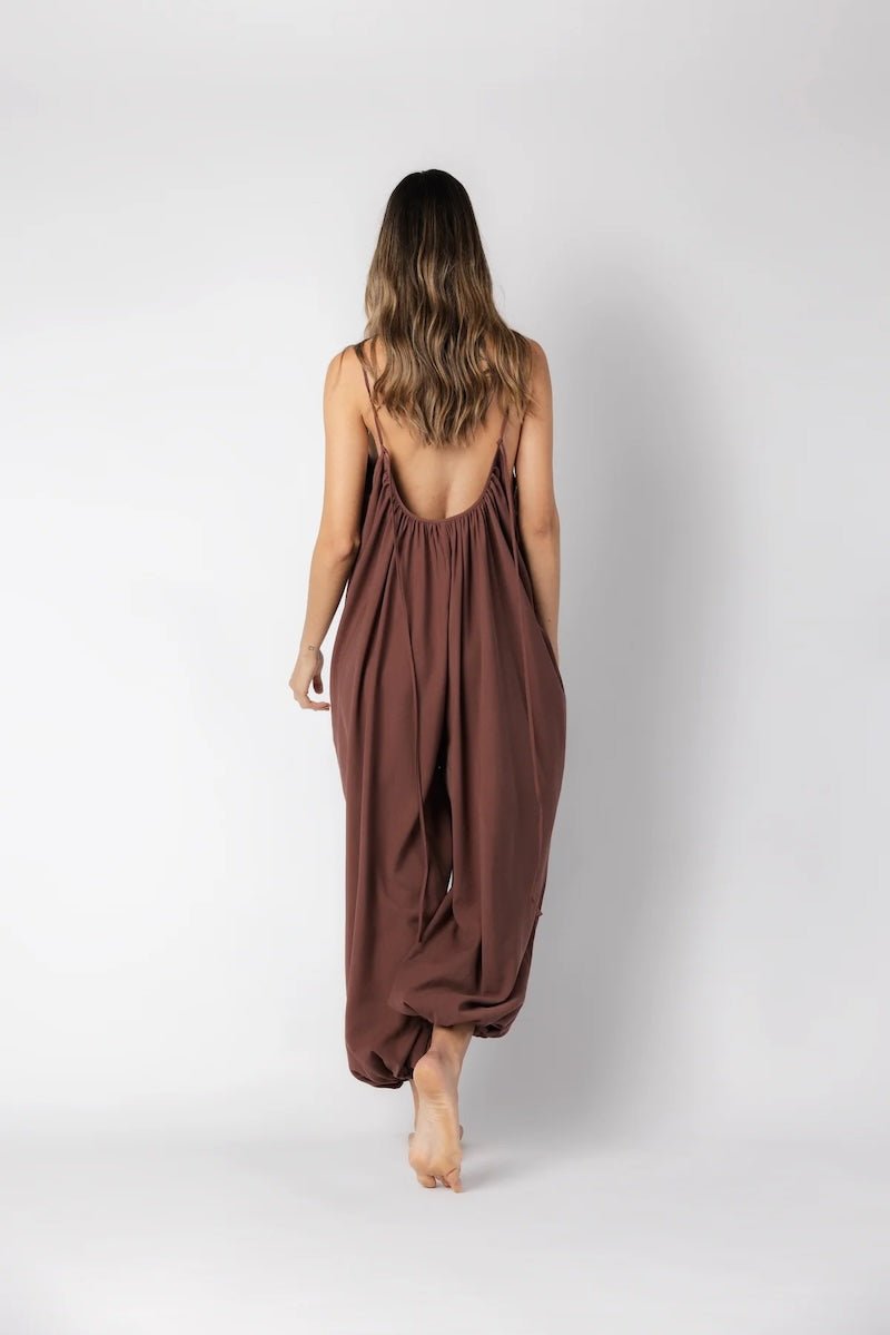 Model wearing a brown jumpsuit. She's standing behind a white background. The purpose of this image is to highlight the jumpsuit, which is available at EVAMAIA boutique in Los Angeles.