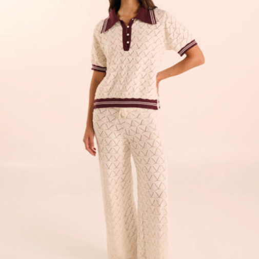 A model wearing a matching two-piece knit lounge set featuring a short-sleeve polo shirt and wide-leg trousers in an off-white textured pattern with dark burgundy trim.