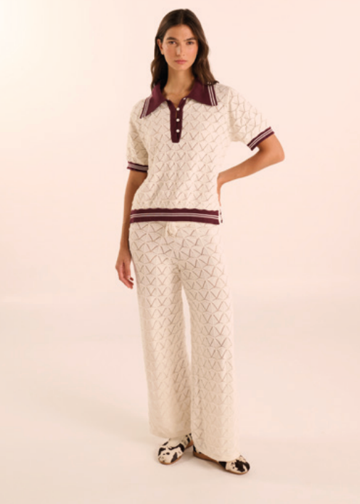 A model wearing a matching two-piece knit lounge set featuring a short-sleeve polo shirt and wide-leg trousers in an off-white textured pattern with dark burgundy trim.