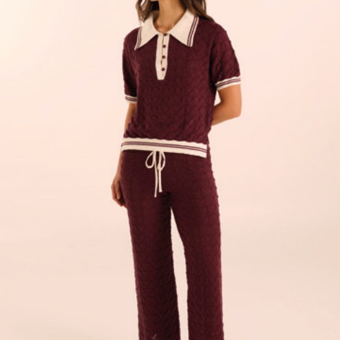 A woman modeling a matching burgundy knit two-piece loungewear set with white accents, including a short-sleeve polo-style top with a white collar and white striped trim, paired with matching drawstring pants, against a plain cream background.