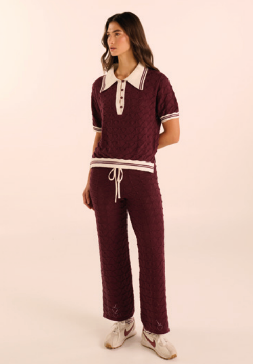 A woman modeling a matching burgundy knit two-piece loungewear set with white accents, including a short-sleeve polo-style top with a white collar and white striped trim, paired with matching drawstring pants, against a plain cream background.